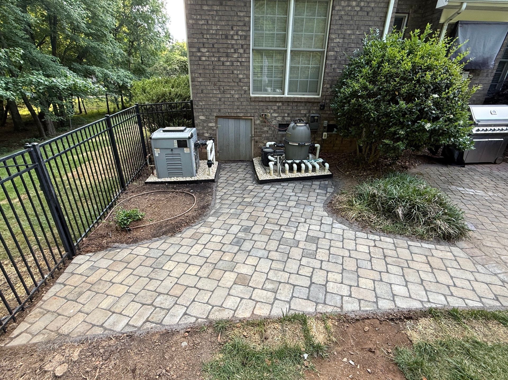 Brick patio with paver path beside a house, black fence, bushes, and utility equipment.
