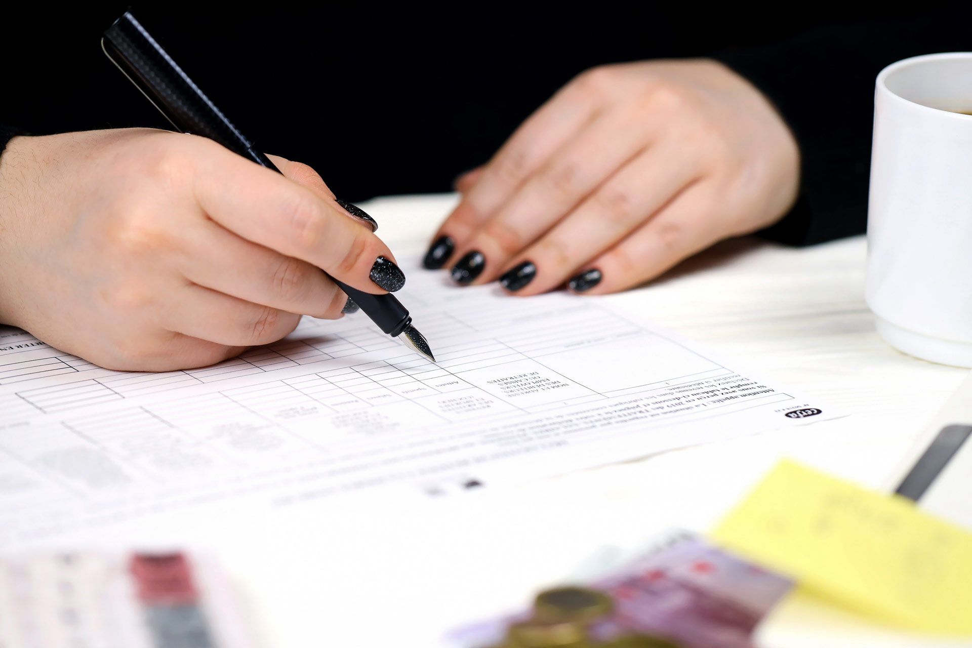 Hands writing on paperwork with a pen, a coffee mug, and money visible on a desk.