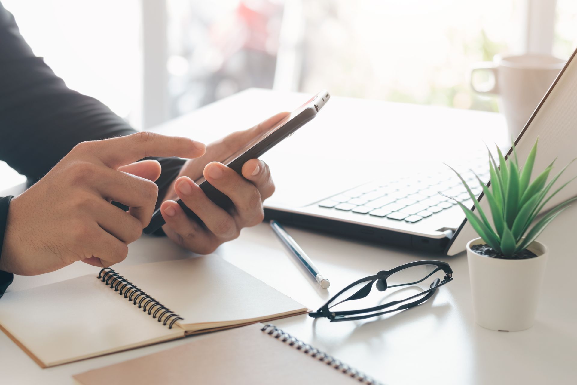 Person using a smartphone at a desk with a laptop, notebook, pen, glasses, and a small plant.