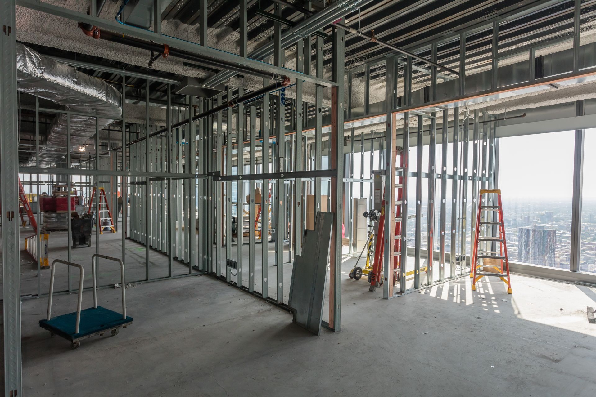 Intérieur d'un bâtiment en construction, charpente métallique et espace ouvert, échelles visibles près d'une fenêtre donnant sur la ville.