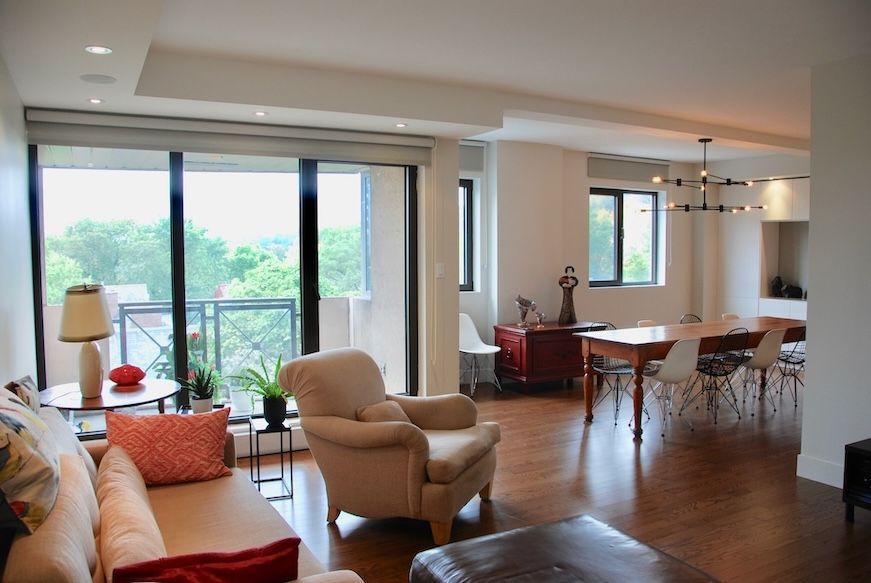 Salon avec une grande fenêtre, parquet et coin repas. Canapé beige, fauteuil et table d'appoint ronde.