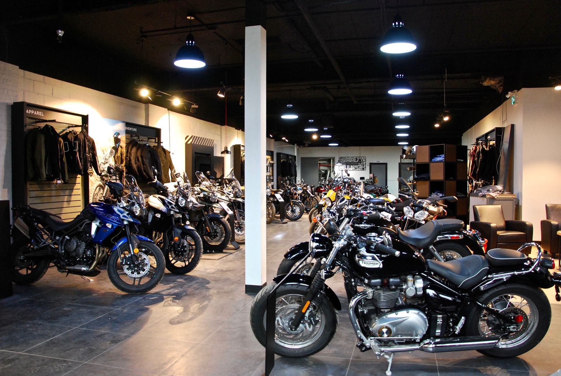 Salle d'exposition de motos. Motos noires et chromées exposées sous des luminaires au plafond. Des blousons en cuir sont accrochés au mur.