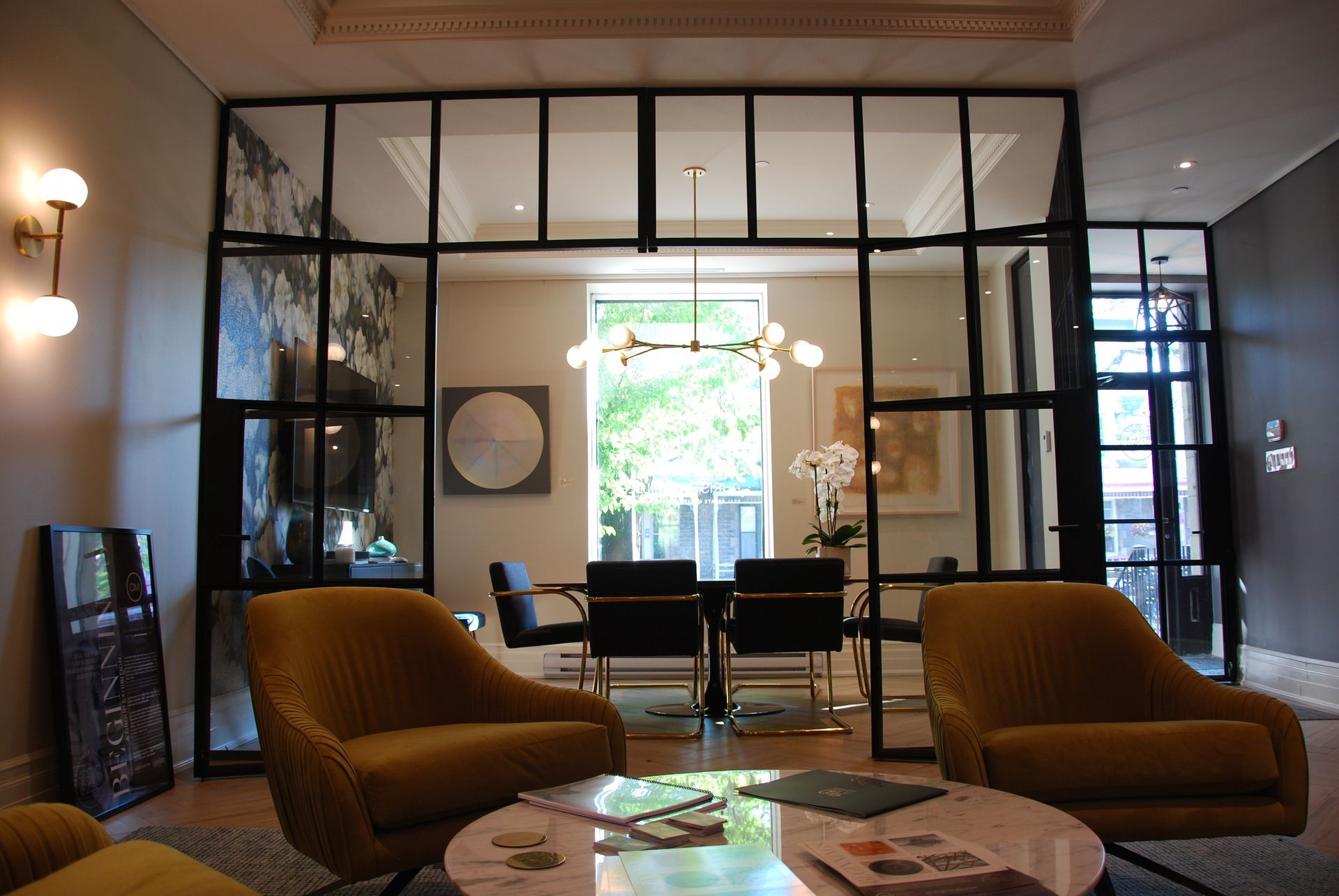 Salon avec fauteuils dorés, salle à manger aux parois de verre, fenêtres à cadre noir et table basse ronde.
