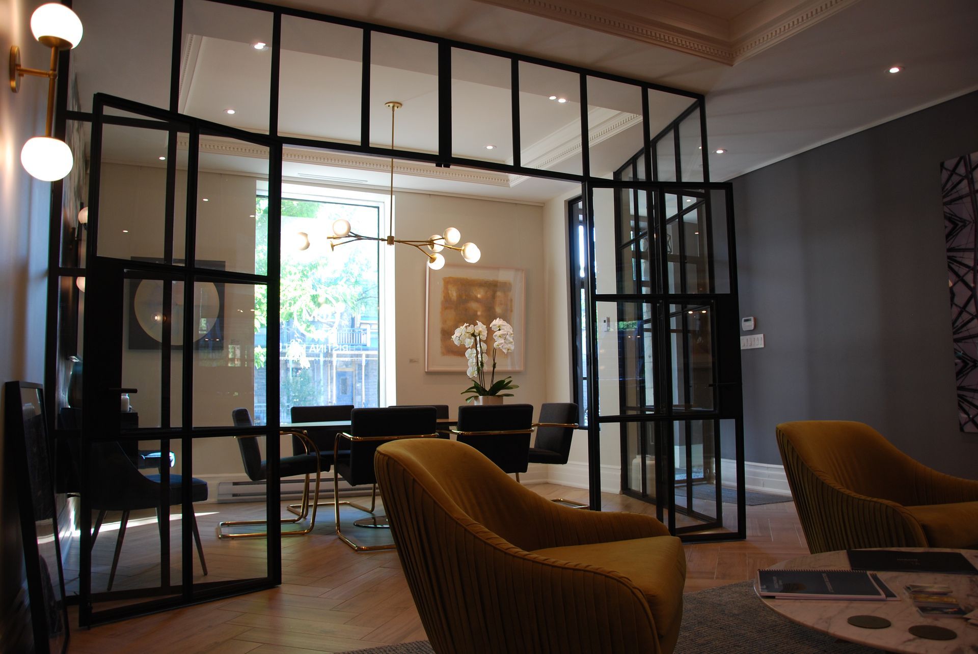 Pièce avec portes vitrées à cadre noir, séparant les espaces salon et salle à manger ; chaises jaunes, table noire, murs blancs.