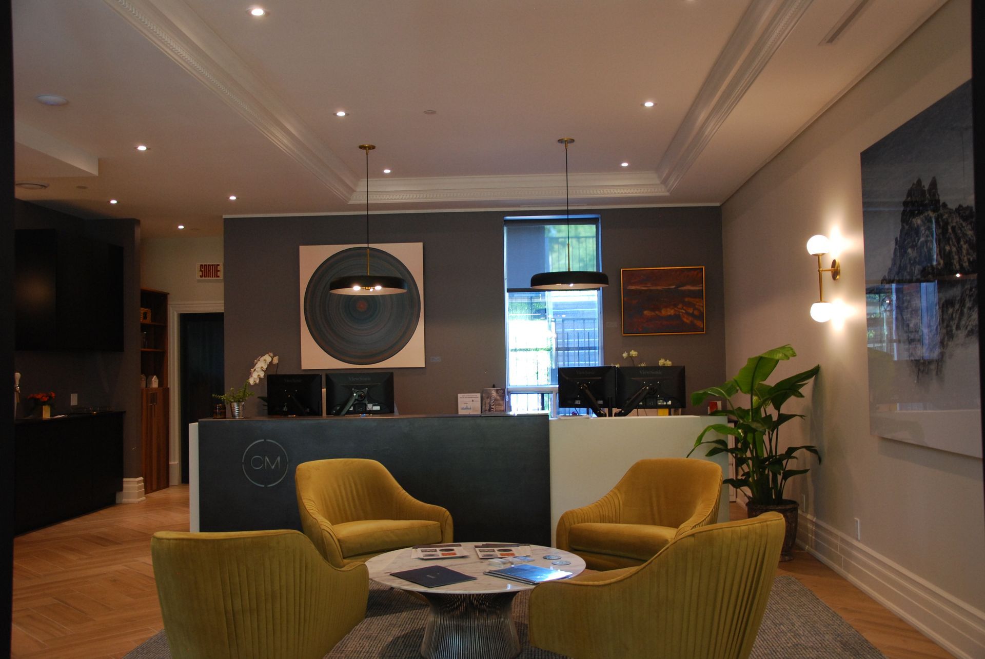 Un espace d'accueil moderne avec des chaises jaune moutarde, une table basse ronde et un comptoir d'accueil gris orné d'œuvres d'art.