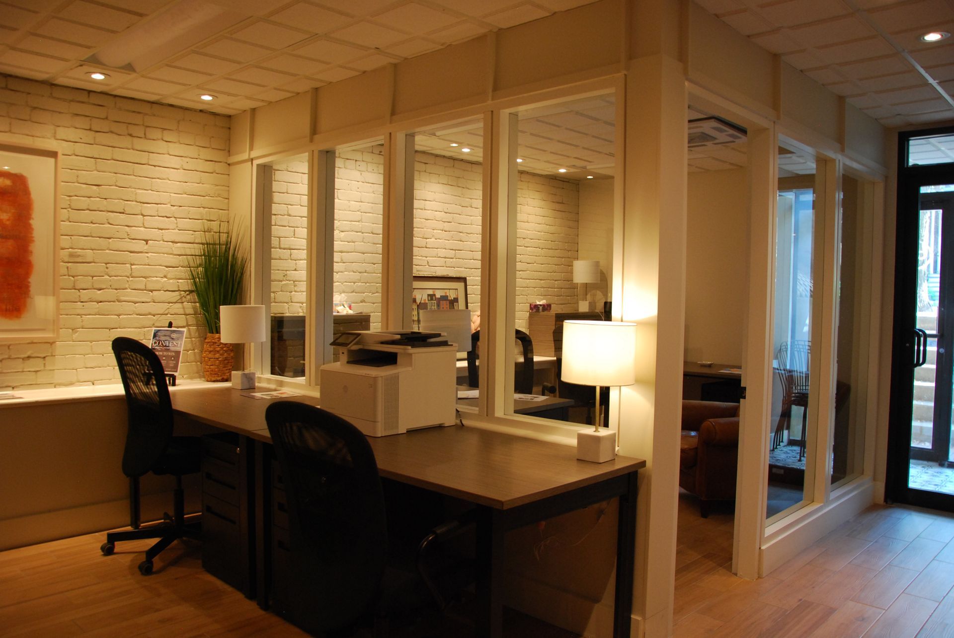 Espace de bureaux avec bureaux, chaises et cloisons vitrées. Murs beiges, tons neutres et éclairage chaleureux.