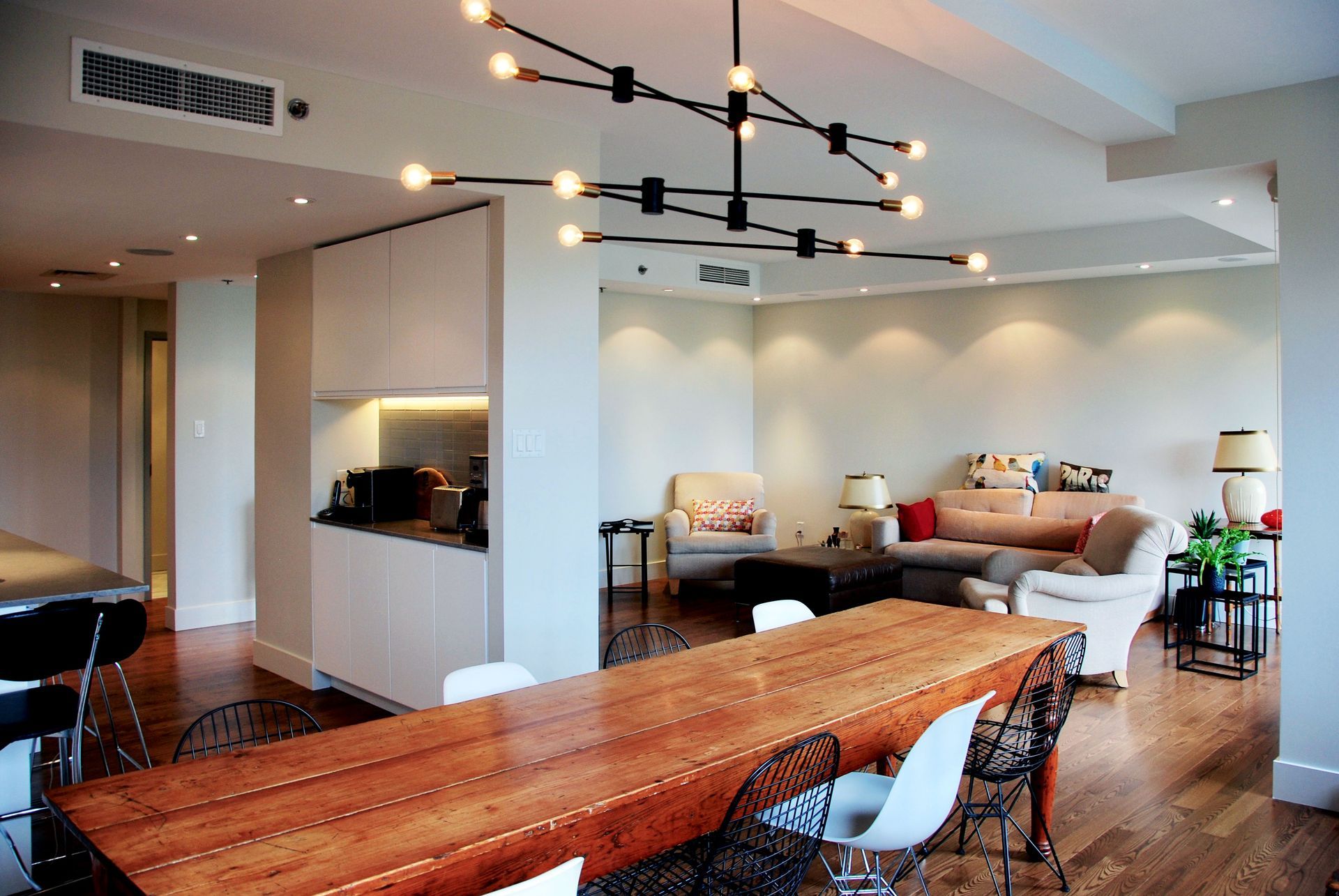 Salle à manger moderne avec une longue table en bois, un lustre et le salon en arrière-plan.