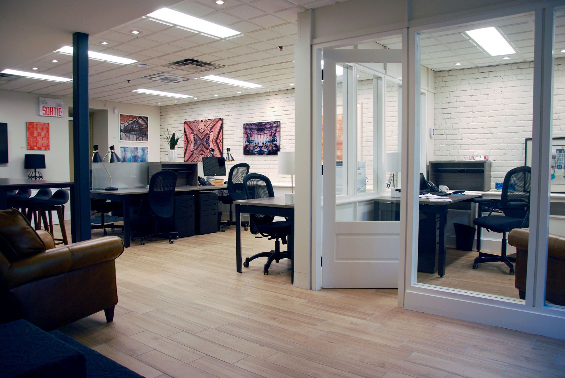Espace de bureaux avec bureaux, chaises et œuvres d'art sur des murs en briques blanches ; un espace de bureaux séparé est visible à travers des portes vitrées.