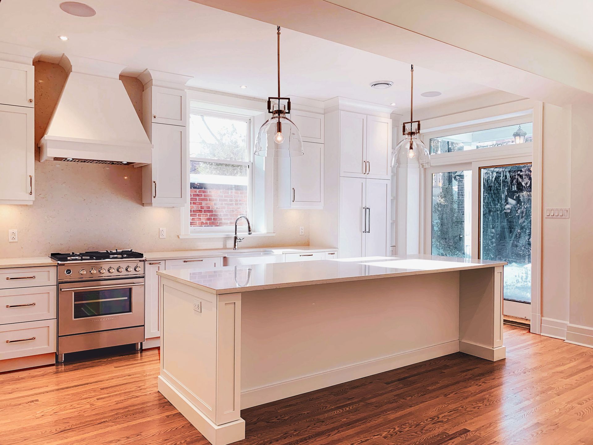 Cuisine lumineuse et blanche avec îlot central, électroménager en inox et parquet. Une fenêtre et une porte vitrée laissent entrer la lumière naturelle.