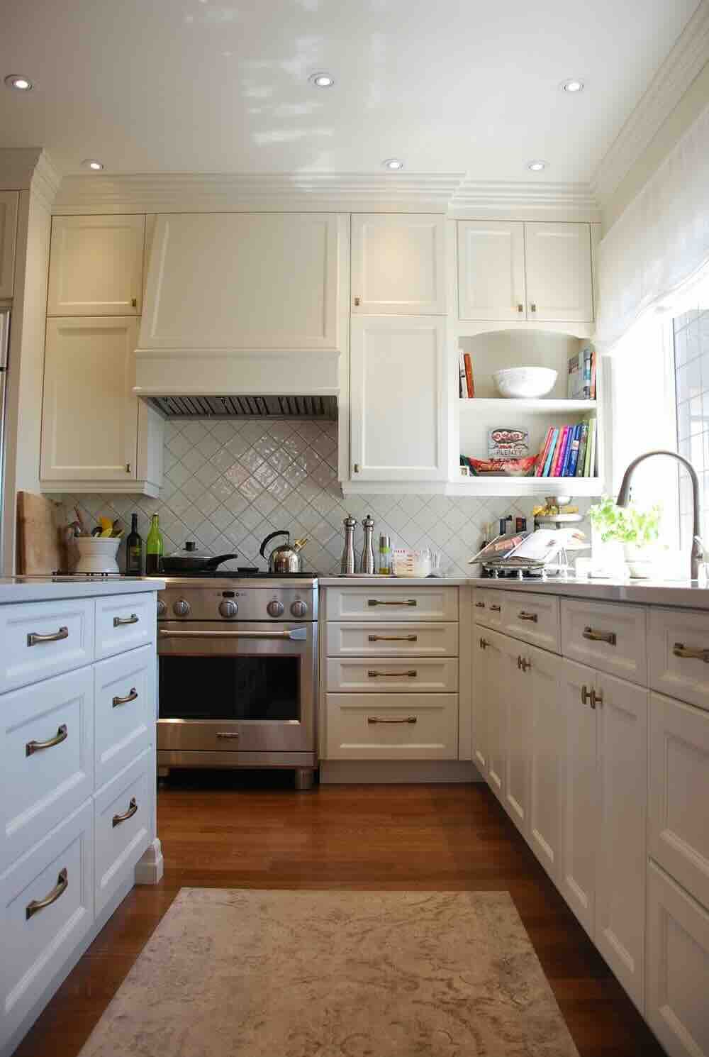 Cuisine blanche avec four en acier inoxydable, armoires blanches, parquet et tapis à motifs.