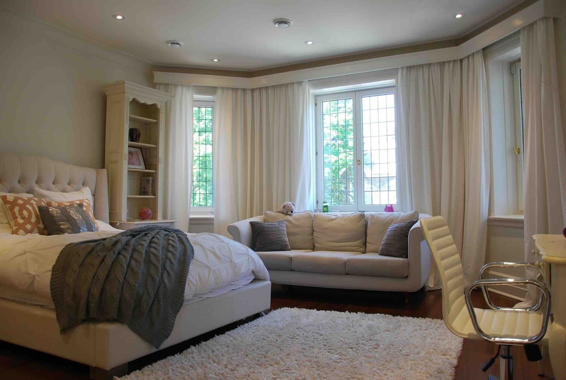 Chambre avec lit, canapé, fenêtres et rideaux blancs. Parquet clair, murs blancs et tapis blanc moelleux.