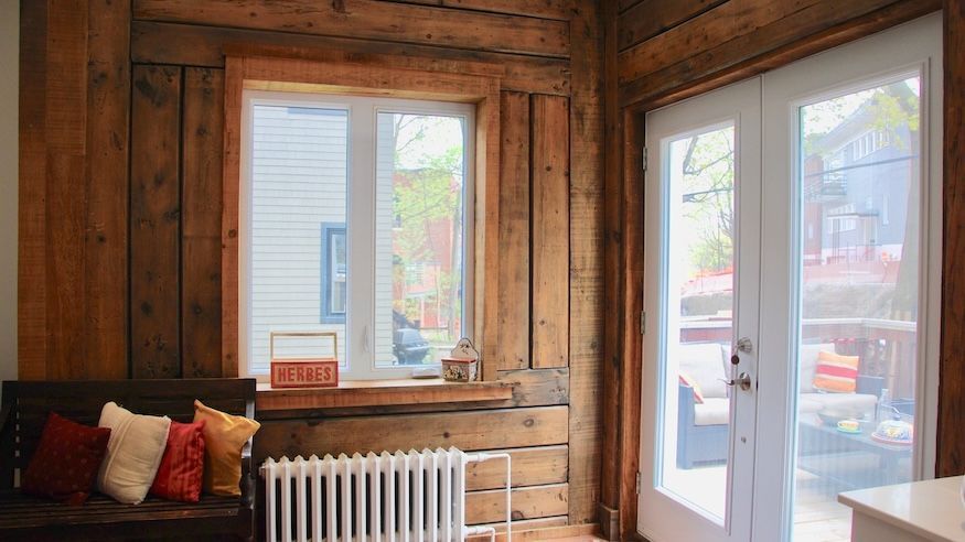 Pièce aux murs en bois avec une fenêtre, un radiateur blanc, une banquette avec des coussins et des portes-fenêtres donnant sur une terrasse extérieure.