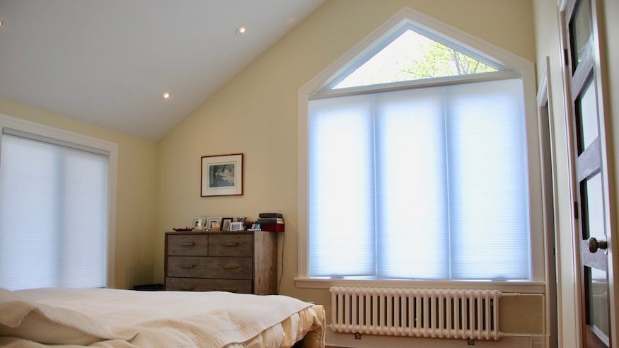 Chambre avec grande fenêtre triangulaire, stores blancs, murs beiges, radiateur et commode en bois.