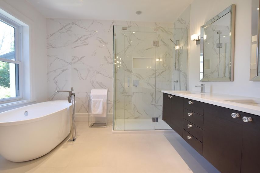 Salle de bains moderne avec baignoire îlot blanche, douche en verre, mur en marbre et meuble-lavabo en bois foncé.
