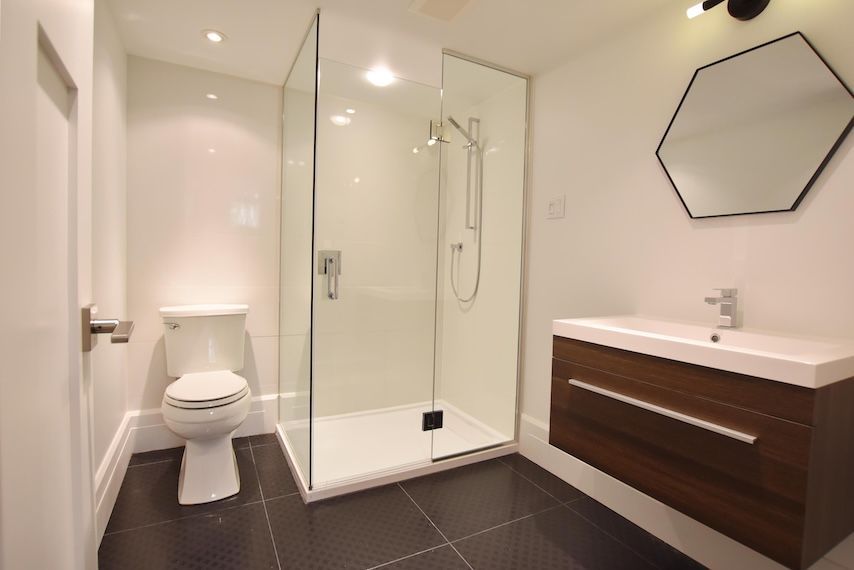 Salle de bains moderne : murs blancs, sol foncé, douche en verre, meuble-lavabo en bois, miroir hexagonal.
