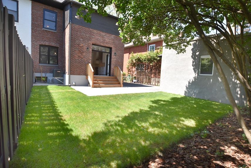Jardin avec pelouse verte, terrasse, maison en briques et clôture en bois. Ensoleillé avec des zones ombragées.