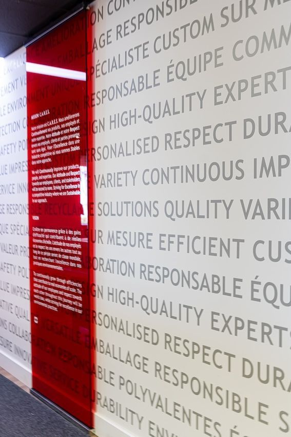 Panneau de verre rouge avec texte, placé à côté d'un mur où se répètent des mots gris, mettant en avant les valeurs de l'entreprise.