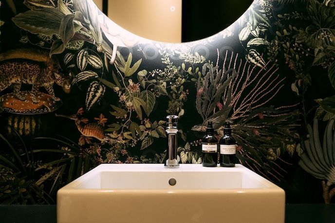 Un lavabo avec un miroir rond, du papier peint à motifs botaniques et des bouteilles sombres.
