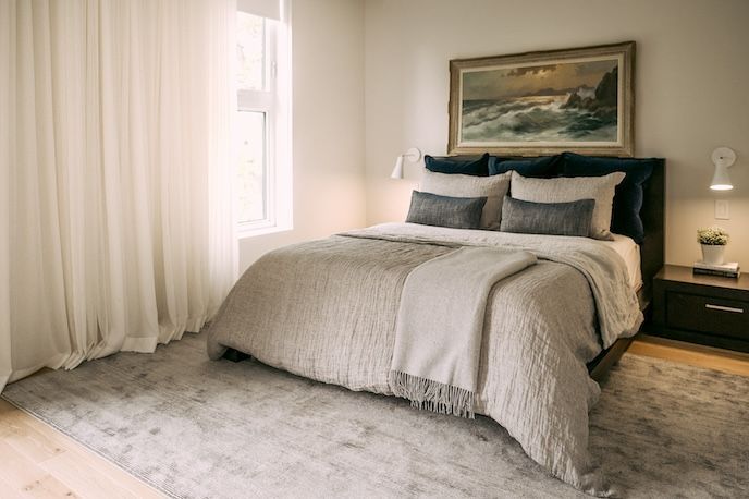 Chambre avec lit, tableaux, voilages, tapis et table de chevet. Couleurs neutres ; espace calme et lumineux.