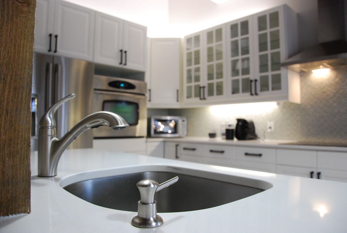 Cuisine blanche moderne avec appareils électroménagers en acier inoxydable, évier au premier plan et armoires avec poignées noires.