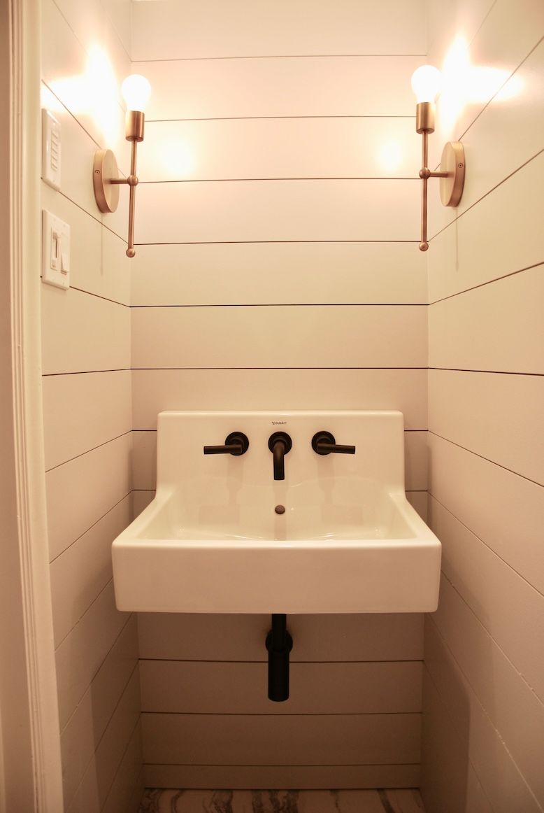 Lavabo mural blanc avec robinetterie noire dans une petite salle de bains. Des appliques encadrent le miroir.