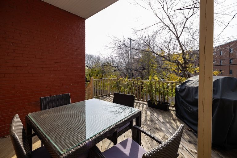 Terrasse avec table en verre, chaises en osier, barbecue et vue sur les arbres. Mur en briques rouges et terrasse en bois.