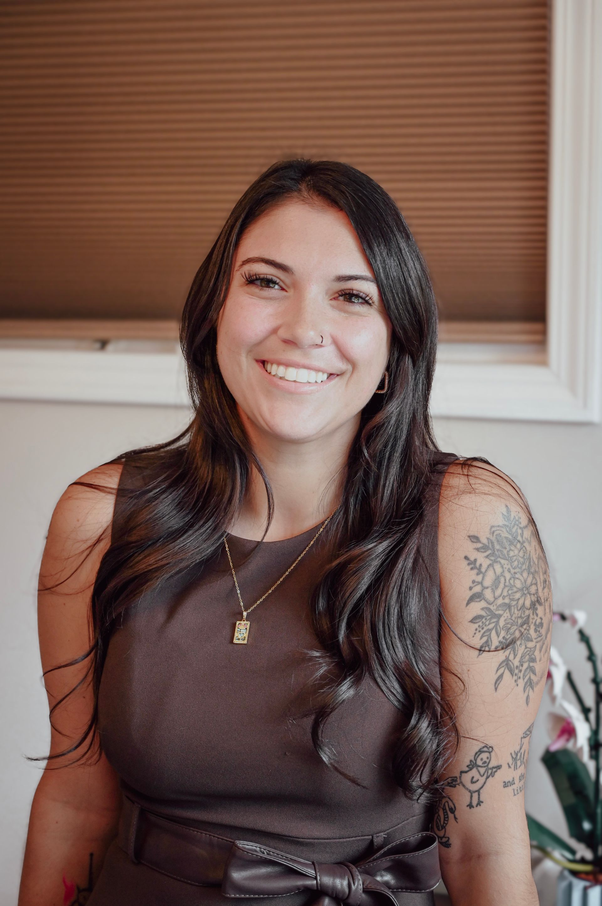 Smiling NC Certified Paralegal with long wavy dark hair, wearing a brown dress and gold necklace, showing floral and artistic tattoos on her arm, posed indoors against a neutral background.