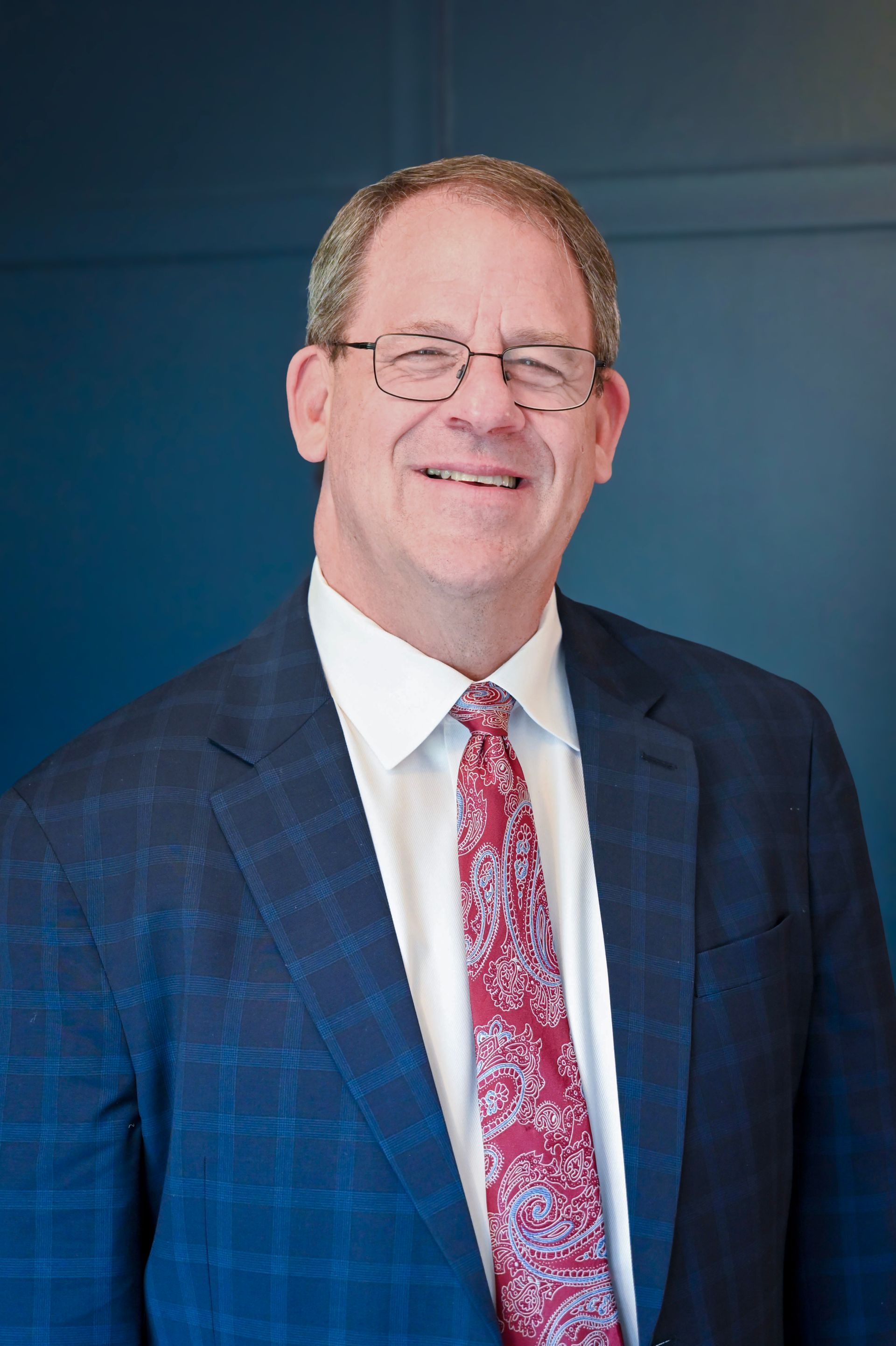 Smiling lawyer in glasses wearing a blue plaid suit jacket, white shirt, and red paisley tie, posed against a plain blue backdrop.