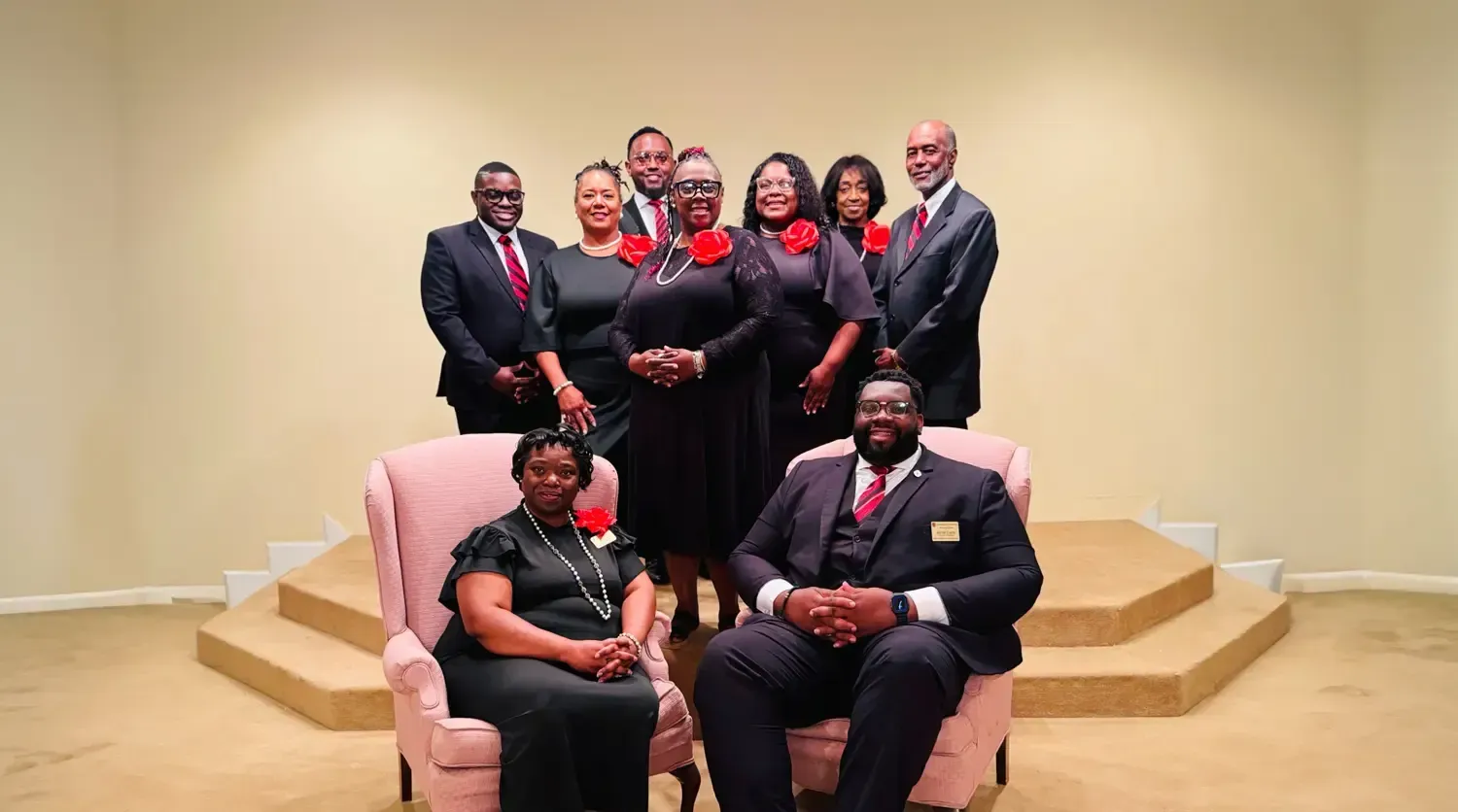 Group portrait of nine people in formal black and red attire on a beige stage with steps and sofa seating