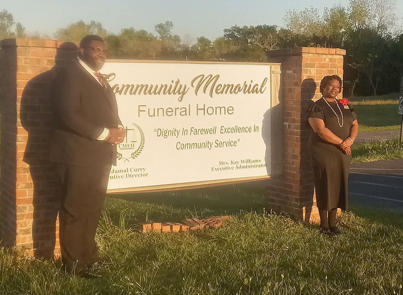 Two people standing beside a Community Memorial Funeral Home sign at sunset.