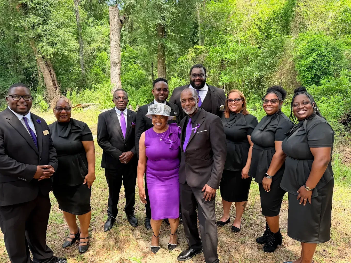 Group of nine people posing outdoors on a wooded path, dressed in formal black and purple attire.