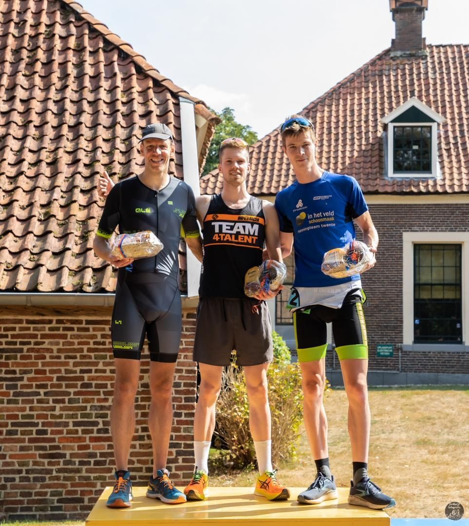 Drie mannen staan ​​op een podium voor een bakstenen gebouw en houden trofeeën vast.