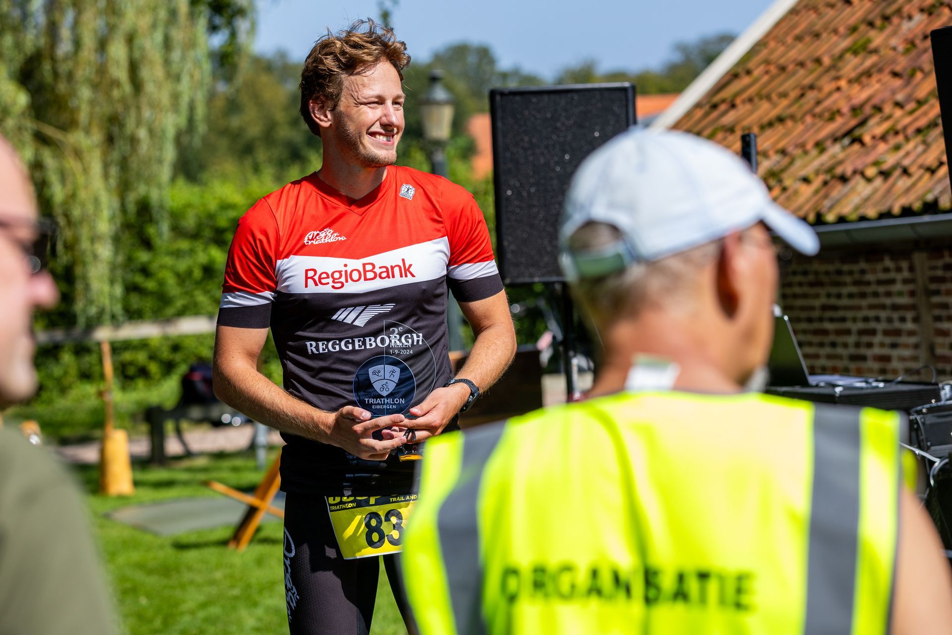 Een man in een rood shirt staat naast een man in een geel vest.