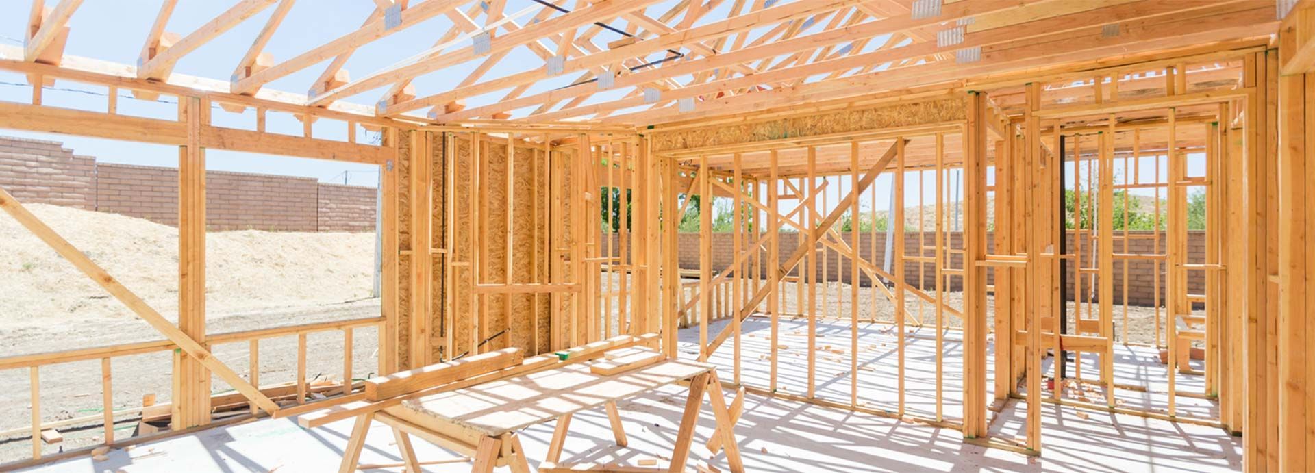 Wood-framed house under construction, showing exposed studs and roof trusses in a bright interior.