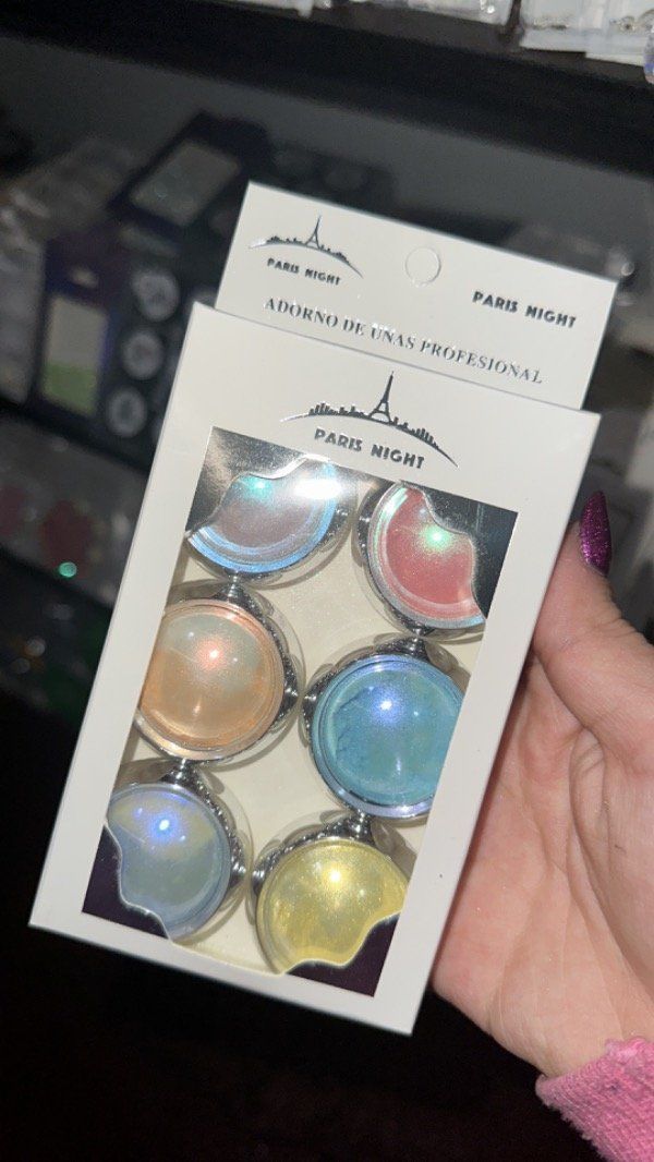 Mano sosteniendo una caja blanca con seis estuches de maquillaje redondos y coloridos, sobre un fondo oscuro.