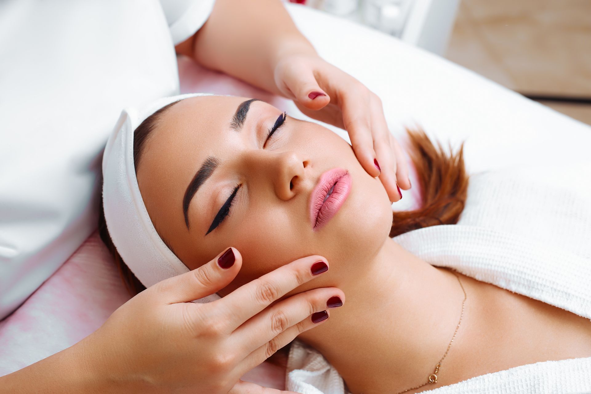 Woman receiving a facial massage at a spa. Hands on cheeks, closed eyes, white towel.