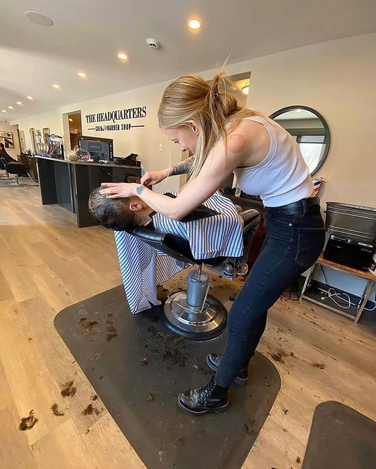 Woman giving a haircut in a salon. The person in the chair is covered with a striped cape. Hair trimmings on the floor.