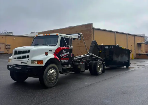 A dump truck is towing a dumpster in a parking lot.