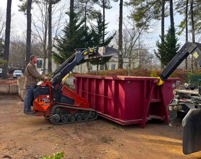A man is driving a tractor next to a large red dumpster.