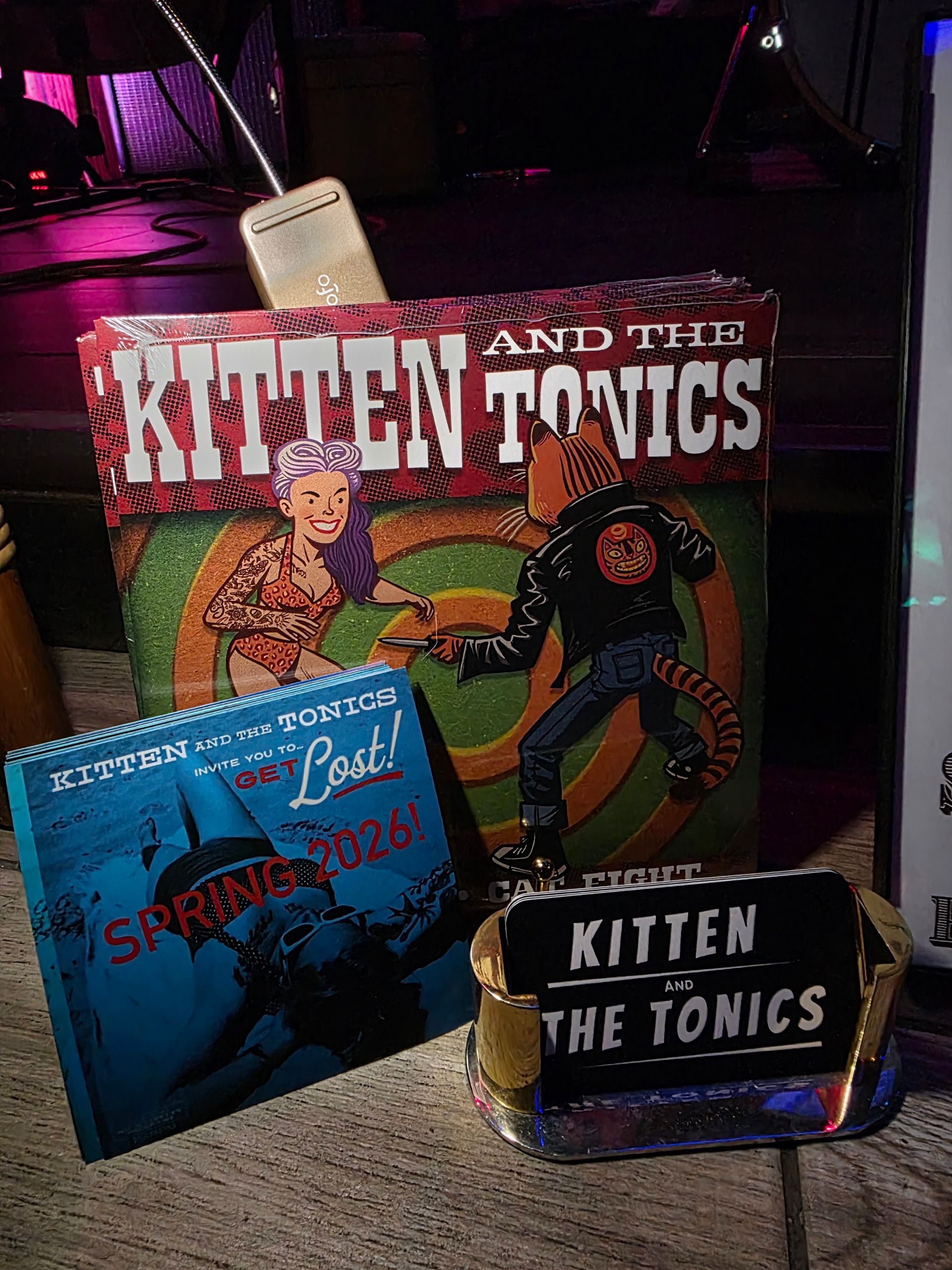 Three pieces of Kitten and the Tonics band merch: a box, a CD, and a button.