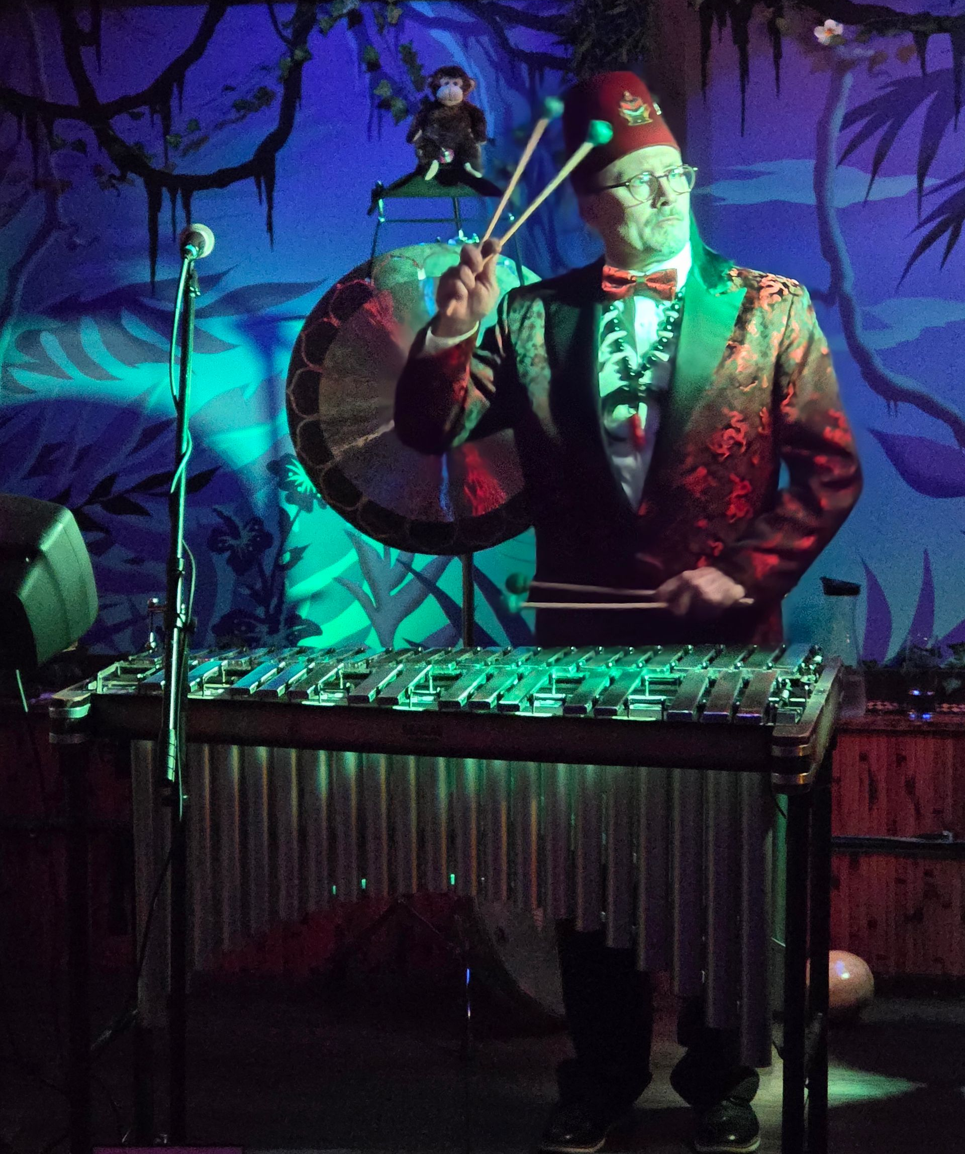 Man in red jacket and fez plays a xylophone with a gong in the background. Green and blue lighting.