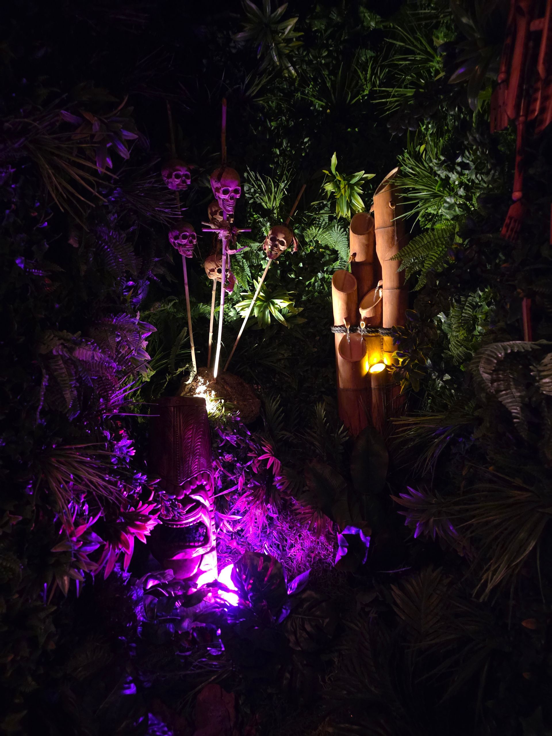 Purple-lit Halloween decorations among green foliage; skulls hang above a bamboo structure with a glowing light.