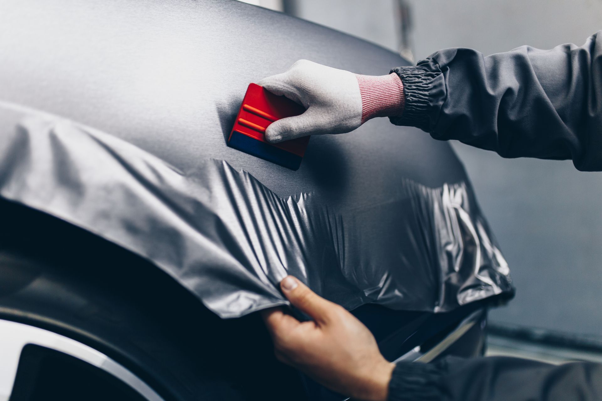 A person uses a red squeegee to smooth a matte gray vinyl wrap onto a car fender.