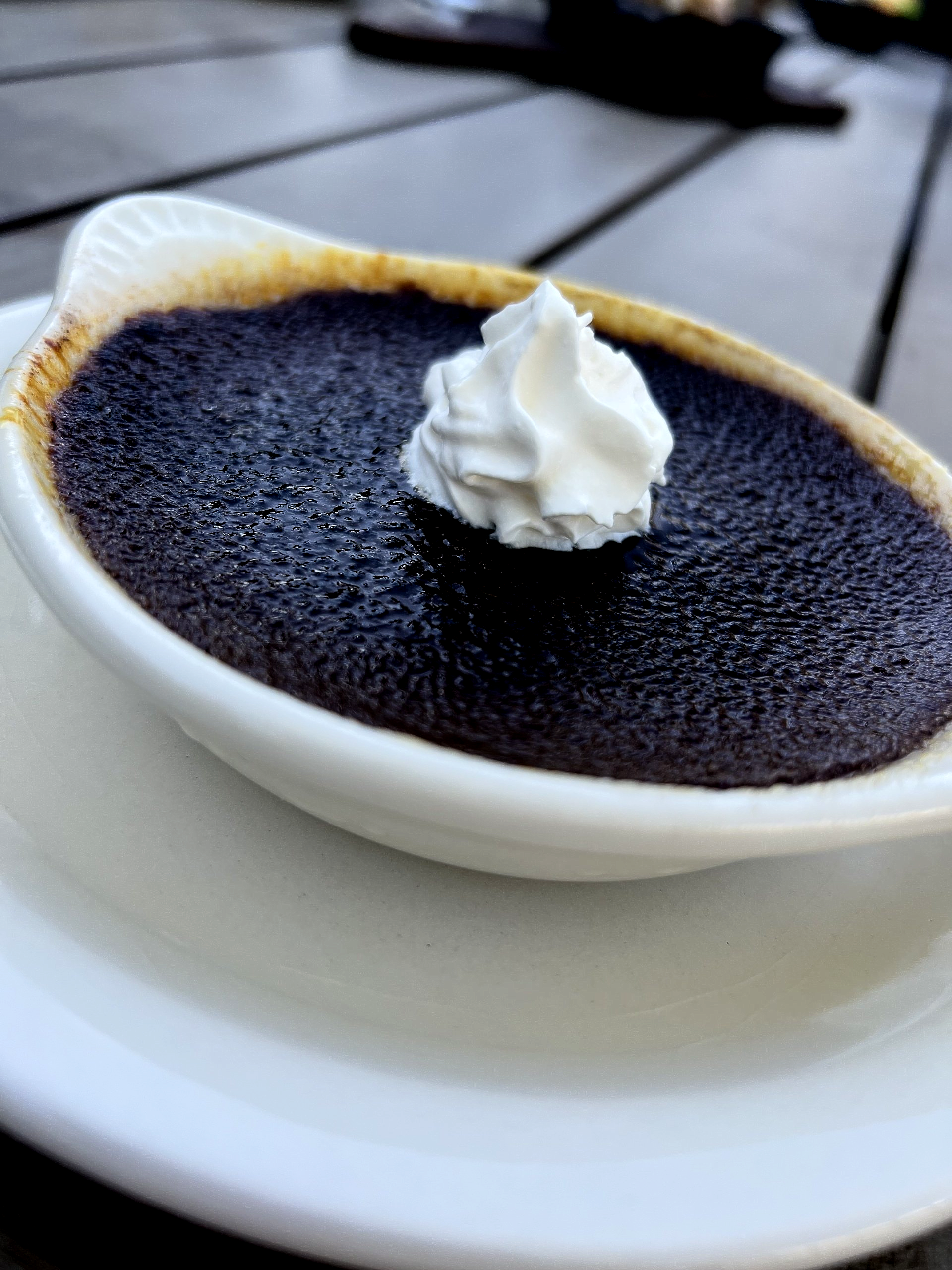 Dark dessert with whipped cream in a white ceramic dish, on a white plate, on a wooden table.