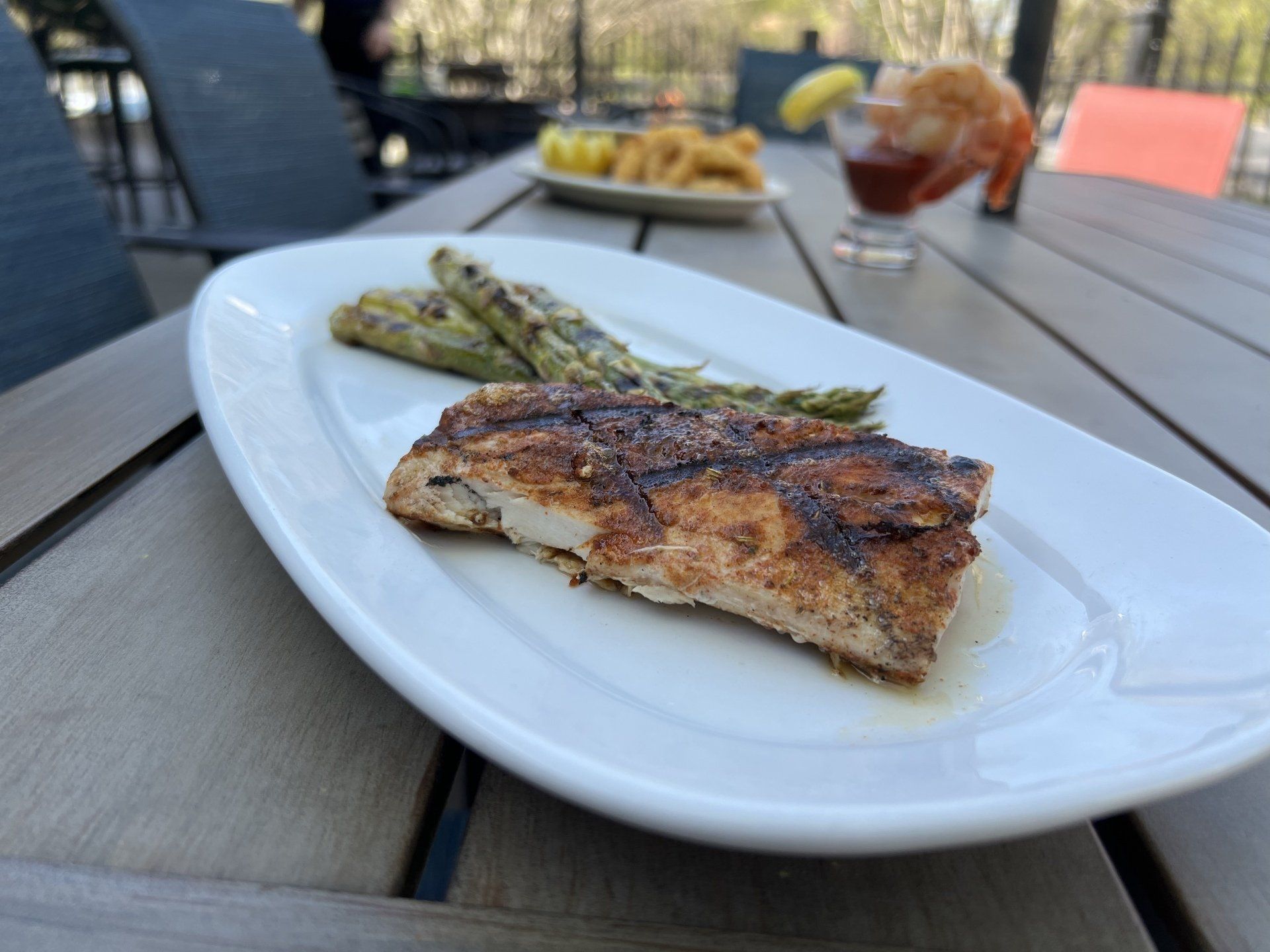 Grilled fish with asparagus on a white plate, served on a wooden table, alongside other dishes.