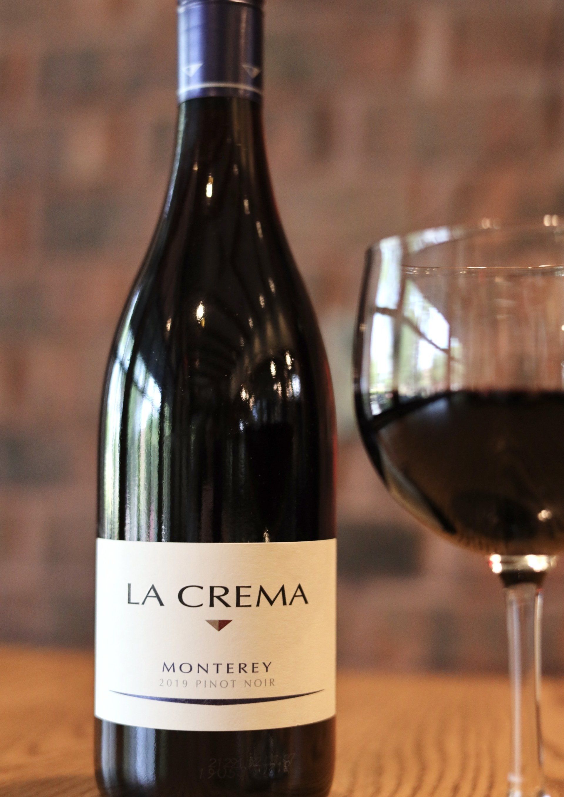 Bottle of La Crema Monterey wine with a glass of red wine, indoors against a brick wall.