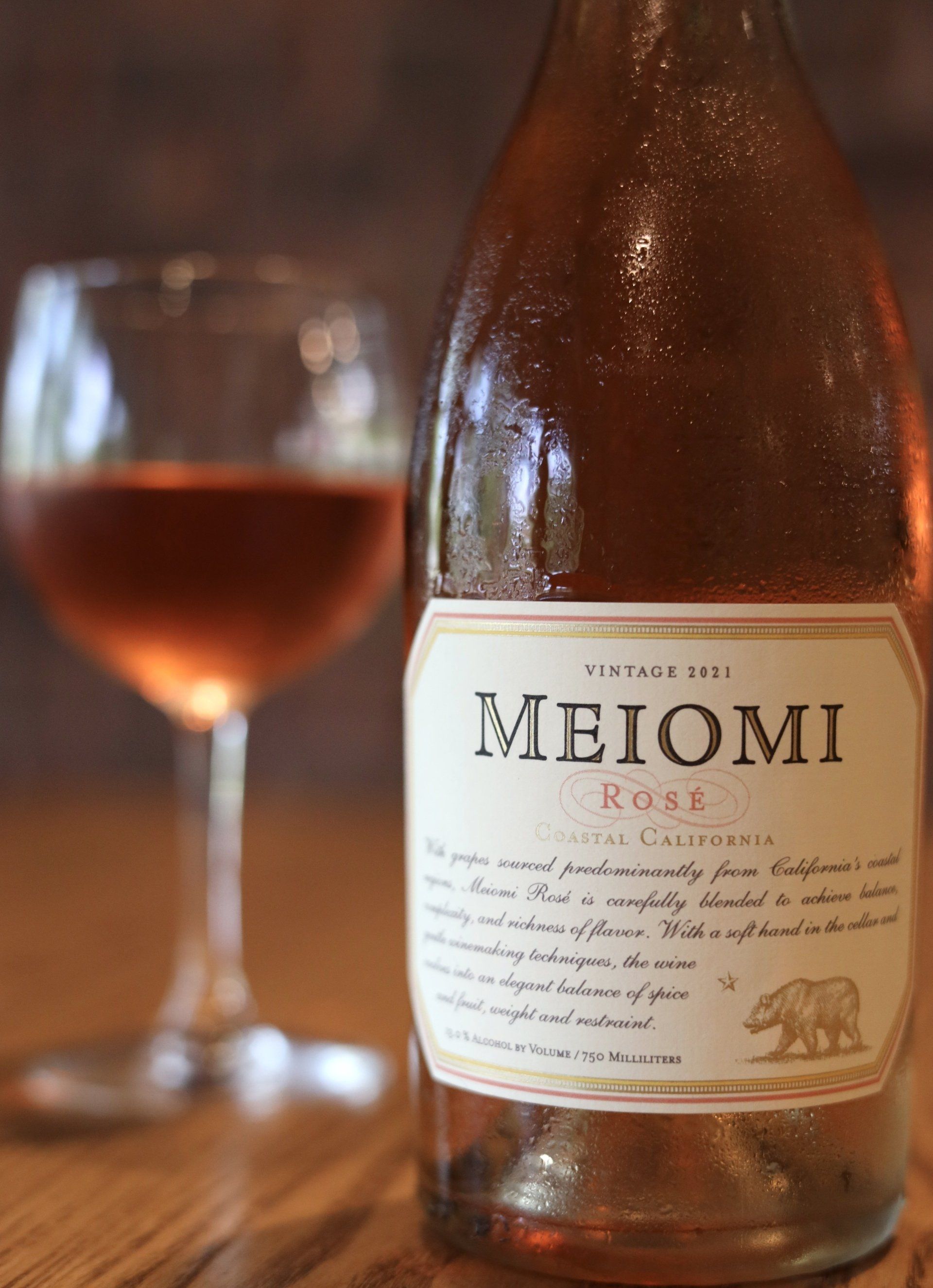 Bottle of Meiomi Rosé wine and glass, both with rose-colored liquid, against a blurred wooden backdrop.