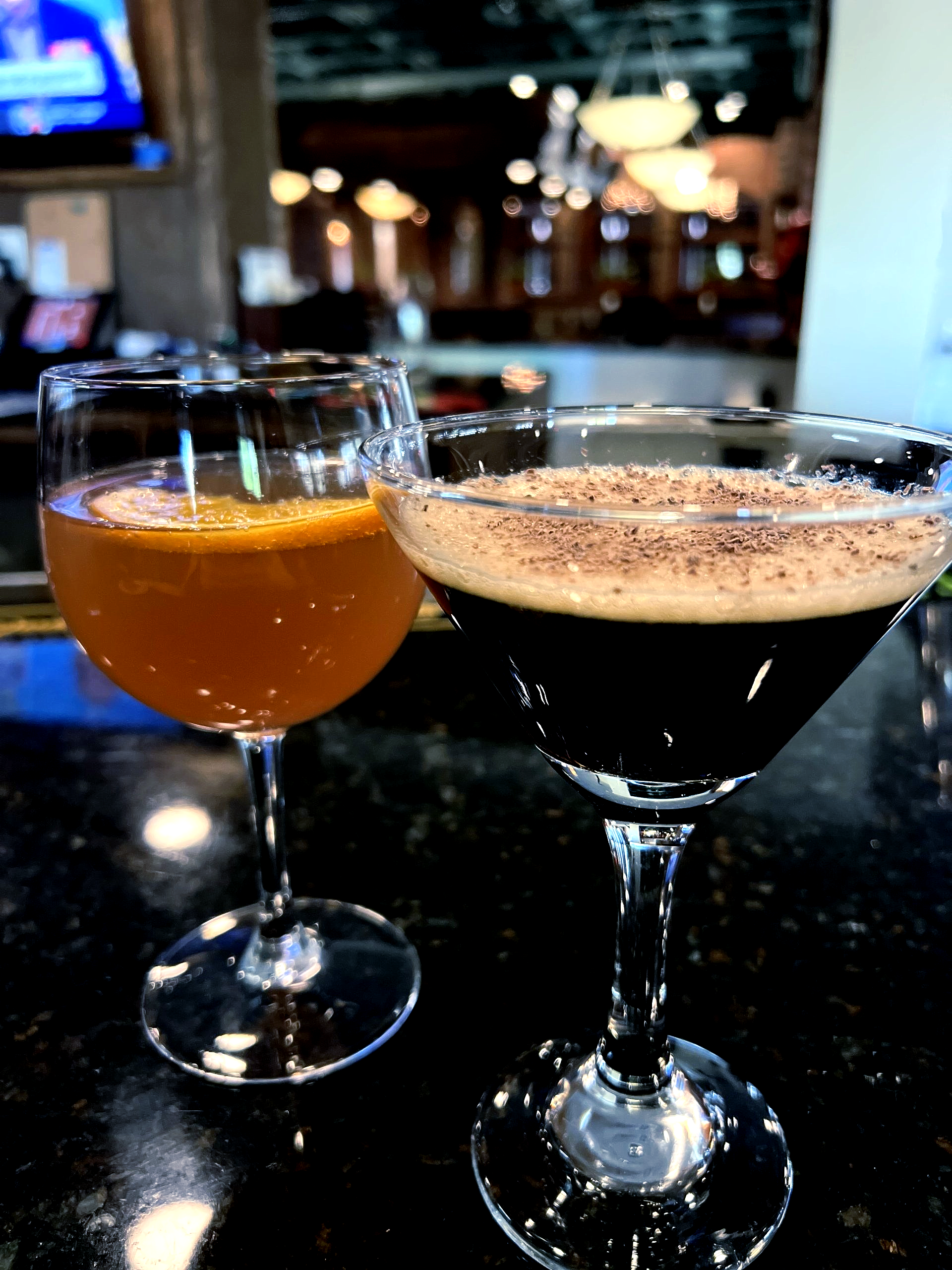 Two cocktails: one amber with orange slice, one dark with cocoa, on a bar.