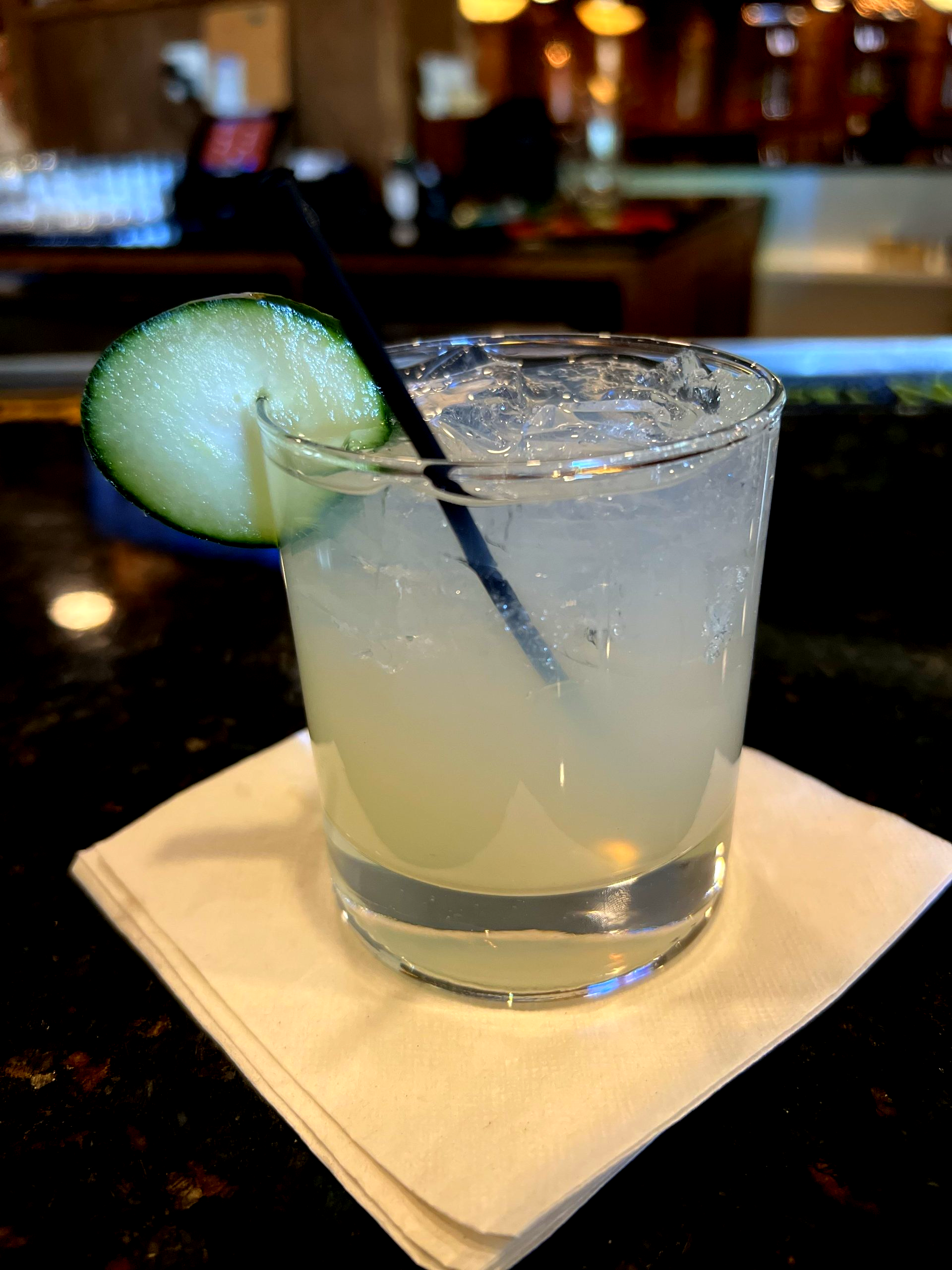 Cocktail in a glass with cucumber slice and straw on a napkin at a bar.