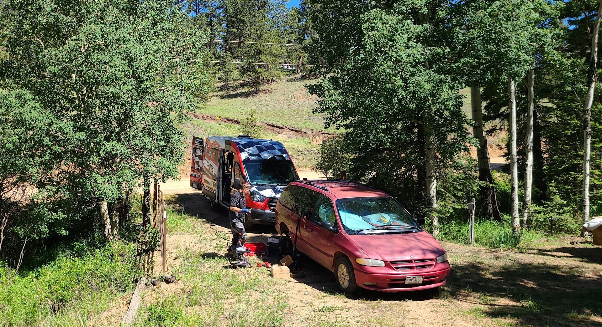 A red minivan being towed by a tow truck on a dirt road in a wooded area on a sunny day. | Elite Mobile Tires and Maintenance